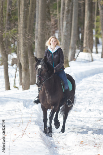 A winter forest. A young blonde woman riding a black horse on snowy ground