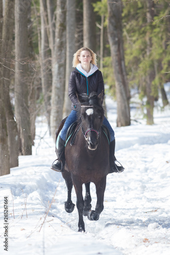 A winter forest. A young woman in warm jacket riding a black horse on snowy ground