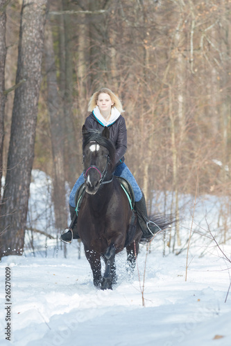 A young blonde woman riding a black horse in the forest