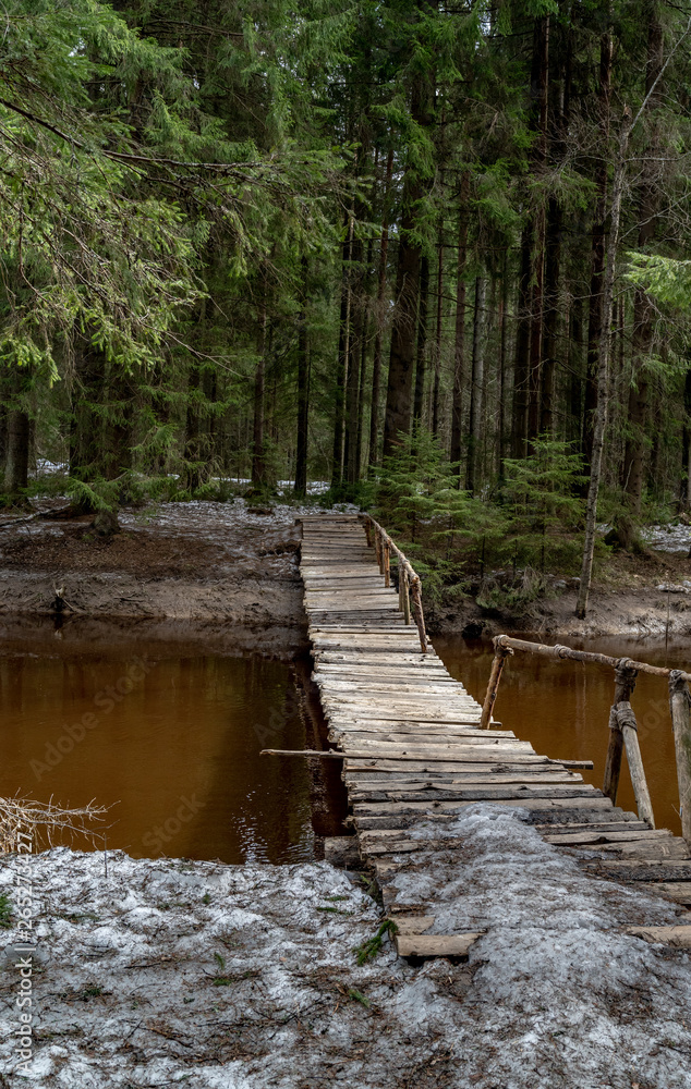 Old wooden bridge over the river in the forest. The ground is covered with a thin layer of snow. Forest in early spring Old wooden bridge over the river in the forest. The ground is covered with a thin layer of snow. Forest in early spring