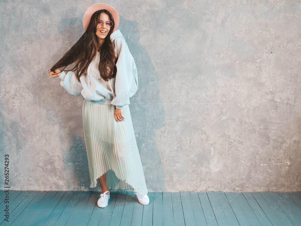 Young beautiful smiling woman looking at camera. Trendy girl in casual summer hoodie and skirt clothes. Funny and positive female posing near gray wall in studio. In hat Young beautiful smiling woman looking at camera. Trendy girl in casual summer hoodie and skirt clothes. Funny and positive female posing near gray wall in studio. In hat