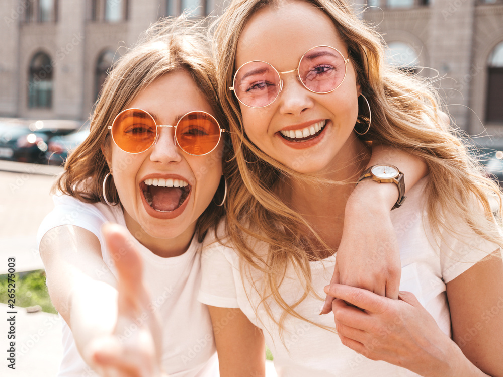 Portrait of two young beautiful blond smiling hipster girls in trendy summer white t-shirt clothes. Sexy carefree women posing on street background. Positive models having fun in sunglasses.Hugging