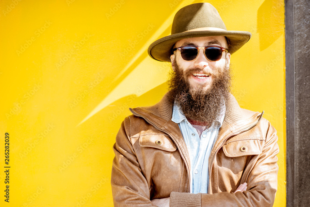 Portrait of a stylish bearded man dressed in jacket and hat on the bright yellow background outdoors Portrait of a stylish bearded man dressed in jacket and hat on the bright yellow background outdoors