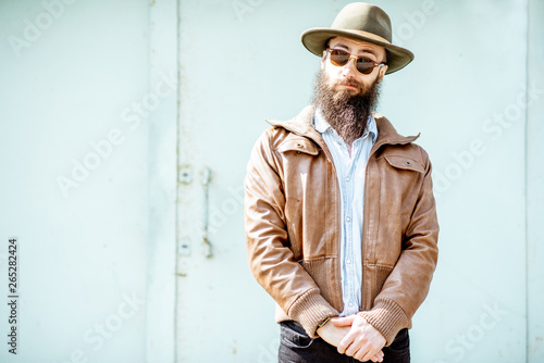 Portrait of a stylish bearded man dressed in jacket and hat on the light turquoise background outdoors
