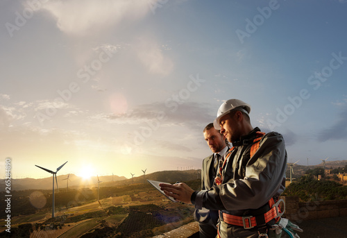 Windmill engineer and businessman planing new ecology project. they standing and looking in tablet. Around wind generators and beautiful landscape