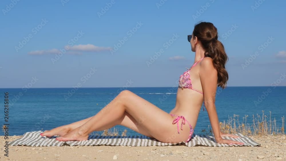 Young woman relaxing on sea shore, sit take sun at height. She wear pink bikini and sunglasses, admire sea from above. Fast motor boat run on water at distance Young woman relaxing on sea shore, sit take sun at height. She wear pink bikini and sunglasses, admire sea from above. Fast motor boat run on water at distance
