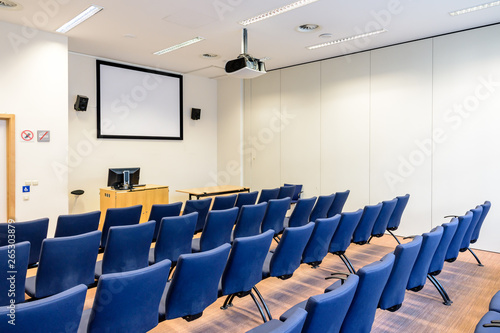 Foto Empty presentation room with rows of blue seats, a white projection screen, a video projector on the ceiling, a computer screen on a desk and white walls