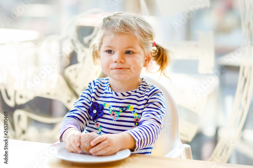 Cute little adorable toddler girl sitting in indoor cafe restaurant. Happy healthy baby child eating bread or sweet scone on sunny day.
