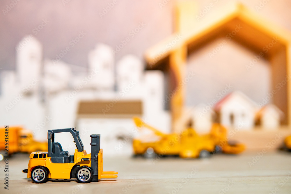 Miniature yellow forklift model on wooden table with blurred house frame and city in the background. Warehouse logistics business or construction industry concept Miniature yellow forklift model on wooden table with blurred house frame and city in the background. Warehouse logistics business or construction industry concept