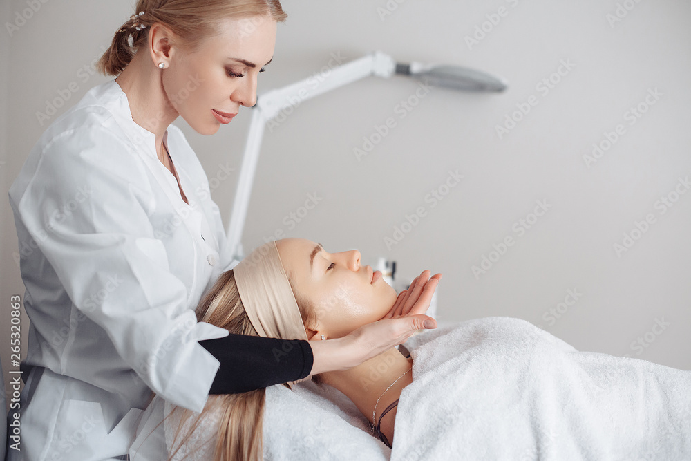 Beautician massage therapist making facial massage to a beautiful female client sitting on a couch in a cosmetology office. Skin care concept Beautician massage therapist making facial massage to a beautiful female client sitting on a couch in a cosmetology office. Skin care concept