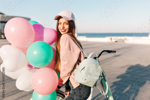 Portrait of adorable smiling brunette girl in trendy pink cap with backpack walking along ocean pier with colorful balloons. Cute young woman with long hair posing at sea wharf after birthday party
