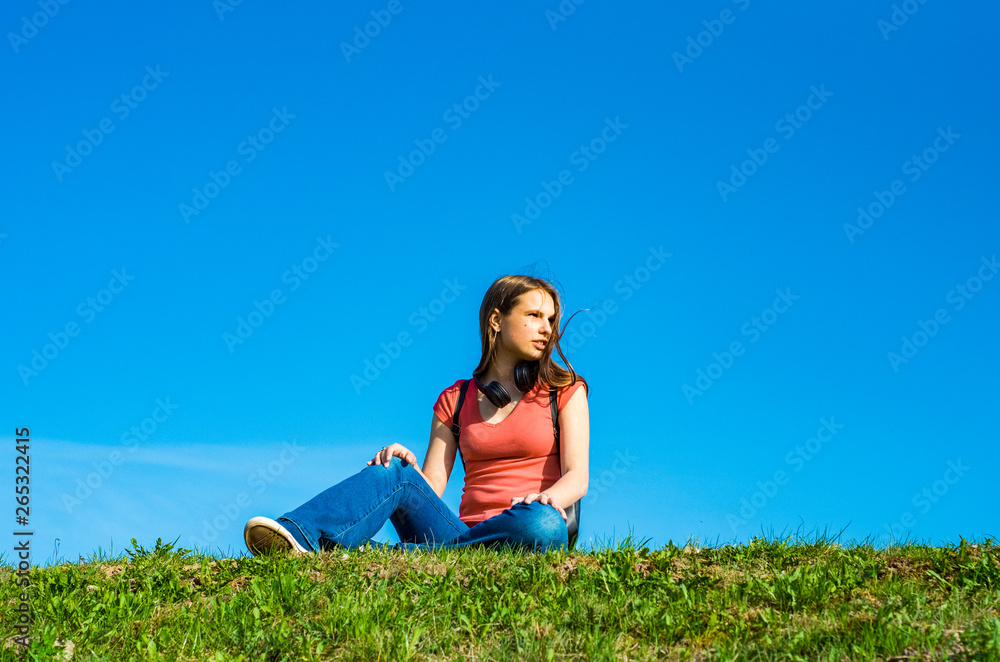 young teenager brunette girl in coral t-shirt with long hair sit on the grass on sky background with copy space young teenager brunette girl in coral t-shirt with long hair sit on the grass on sky background with copy space