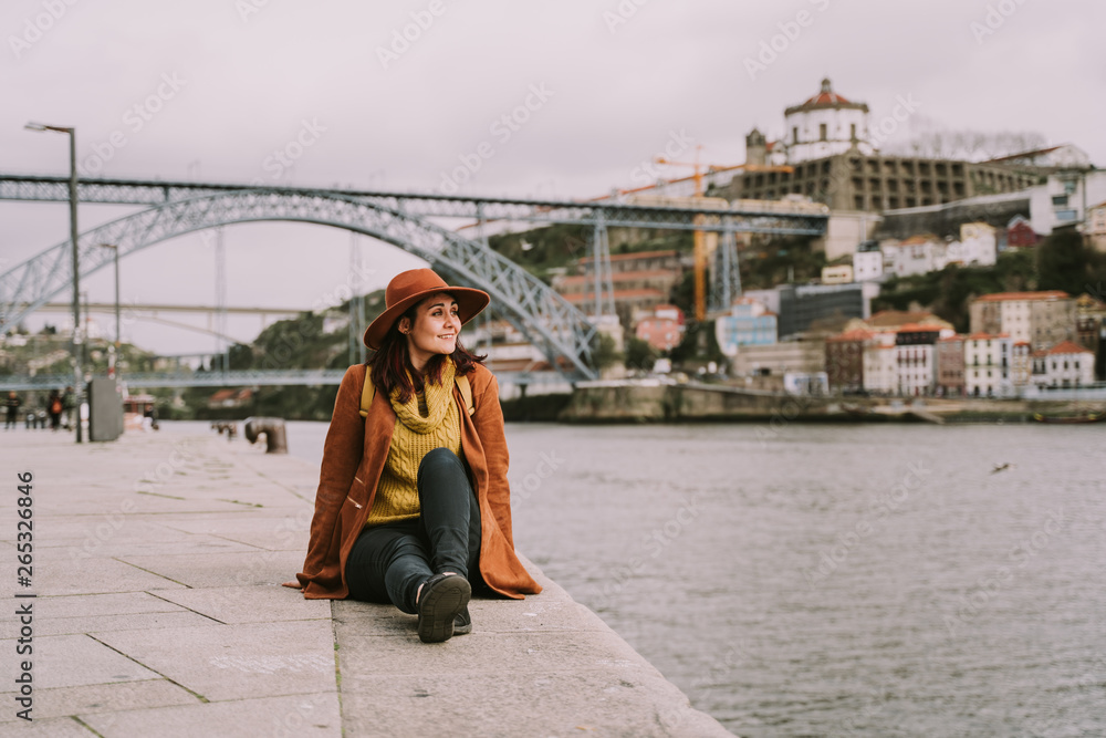 Young traveler woman is enjoying sightseeing in the city of Porto. She is sitting on the riverbank enjoying the landscape and a beautiful sunny day. Travel concept