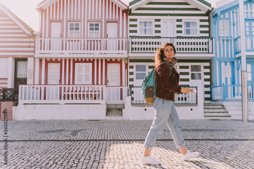 A young beautiful traveler walking by the streets of a Portuguese city. She is wearing jeans, a dark red jacket and a blue backpack. There are colorful houses behind her.