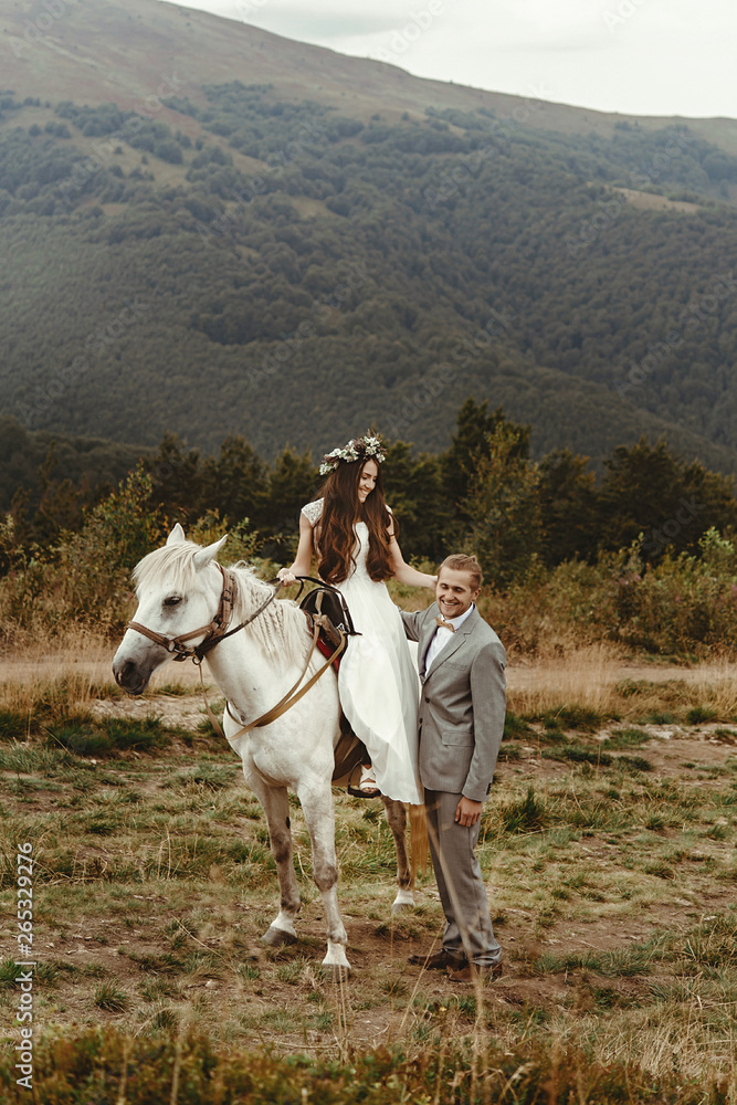 gorgeous bride riding a white horse and stylish groom,  boho wedding couple, luxury ceremony at mountains, space for text
