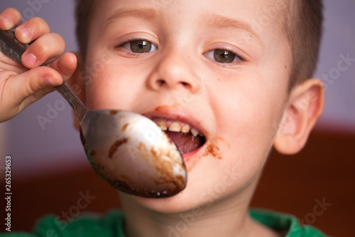 Close up portrait of Cute little boy eating chocolate yogurt at breakfast. Face messy with chocolate and naughty facial expression.