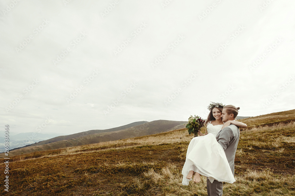 stylish groom carrying happy bride and having fun, boho wedding couple, luxury ceremony at mountains with amazing view, space for text