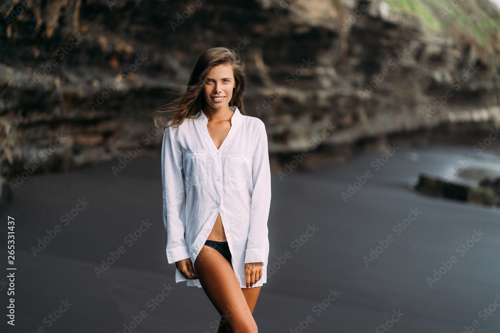 Young sexy girl in white shirt and swimsuit resting on black sand beach. Young sexy girl in white shirt and swimsuit resting on black sand beach.