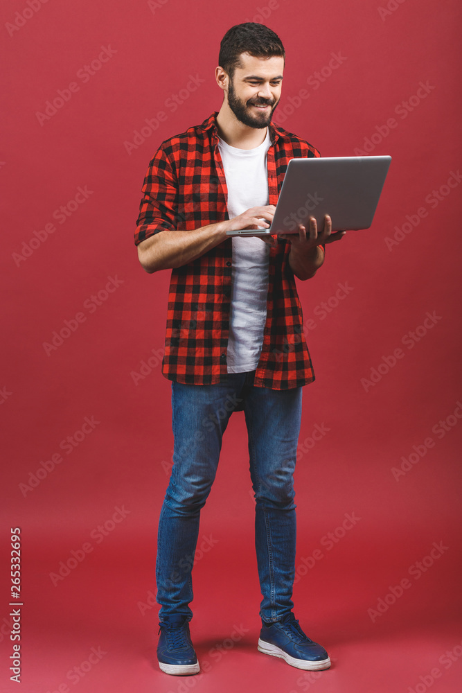 Concentrated on work. Full length portrait of a cheerful young man. Confident handsome man in casual working on laptop while standing against red background. Concentrated on work. Full length portrait of a cheerful young man. Confident handsome man in casual working on laptop while standing against red background.