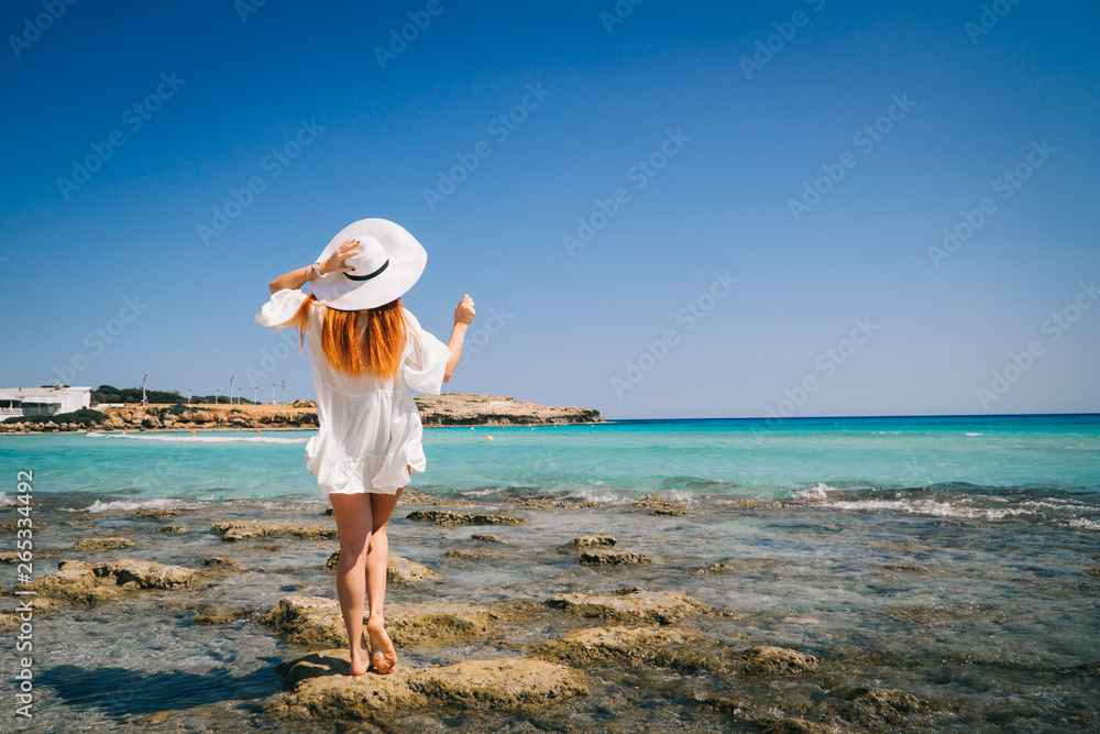 Red-haired female in white dress stands with his back to camera on shores of Mediterranean Sea Red-haired female in white dress stands with his back to camera on shores of Mediterranean Sea