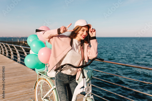 Laughing girl in pink cap coming to sea after bike trip to enjoy beautiful view in morning. Adorable brunette young woman having fun at the ocean and posing with peace sign next to bicycle © Look!