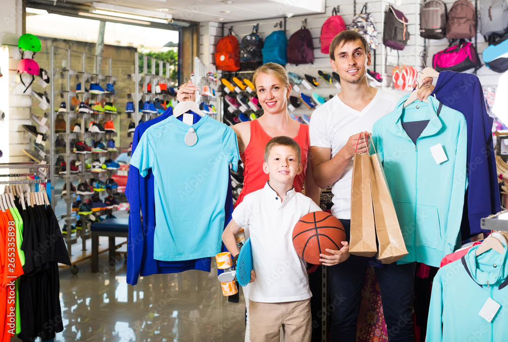Fototapeta premium man and woman with boy choosing t-shirts and other goods in sport shop