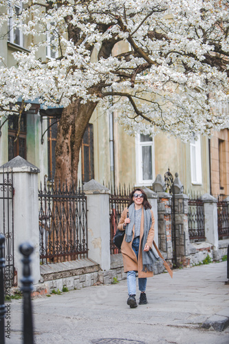 young stylish woman walking by city street in coat blooming tree on background © phpetrunina14