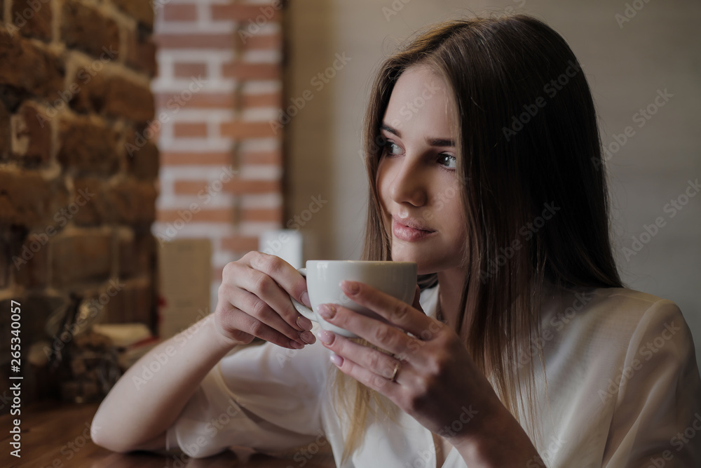Young beautiful brunette girl in a loft style cafe sits at a table by the window, drinks coffee and eats a dessert cake. Young beautiful brunette girl in a loft style cafe sits at a table by the window, drinks coffee and eats a dessert cake.