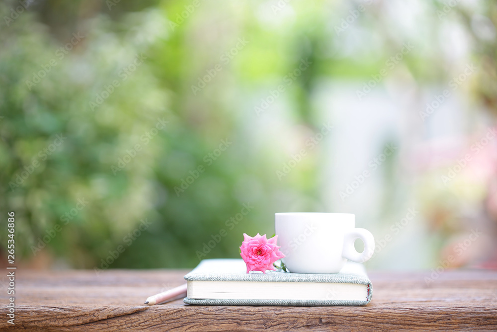 White coffee cup and notebook with pink pygmy rose on wooden table White coffee cup and notebook with pink pygmy rose on wooden table