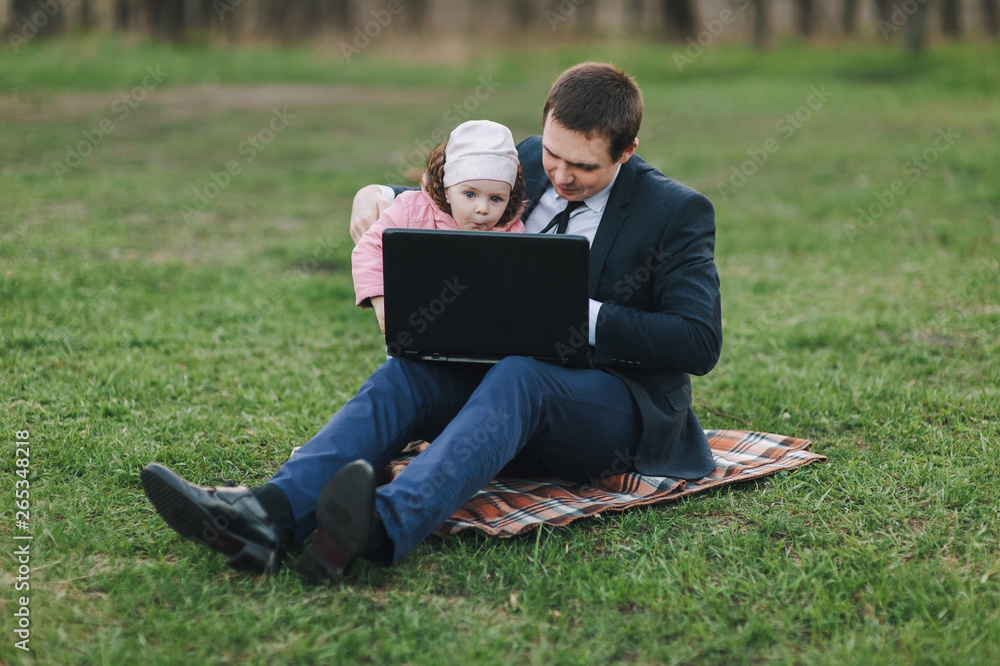 A young businessman in a suit sits in the park on the green grass and watches a movie on a laptop with his beloved child. Computer, lifestyle, work. Father and daughter, business, emotions. A young businessman in a suit sits in the park on the green grass and watches a movie on a laptop with his beloved child. Computer, lifestyle, work. Father and daughter, business, emotions.