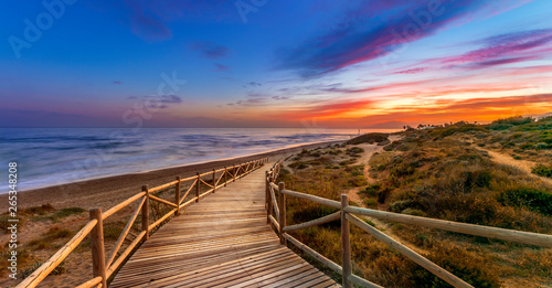Fototapeta Naklejka Na Ścianę i Meble -  View of amazing bright sundown sky over waving sea and wooden path in countryside in Cabopino, Artola dunes. Marbella, Spain