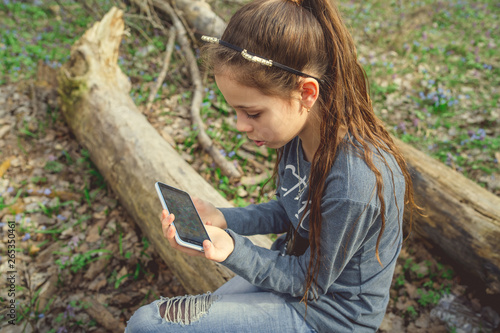Girl sitting on a log in spring forest, uses her smartphone Teenager in outerwear sitting on a fallen log in a rural forest looking at the phone screen