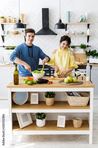 Beautiful couple in kitchen. Handsome man is preparing a salad while his girlfriend is cutting bread, both are smiling and have fun time