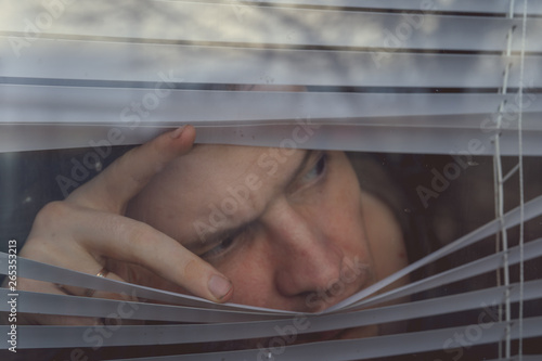 Man watching through window blinds Portrait of young thoughtful male with brown eyes observing through window jalousie