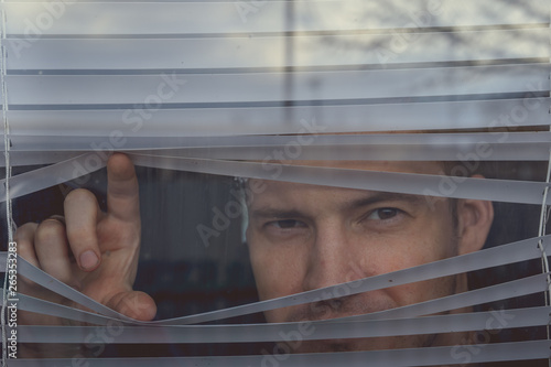 Man watching through window blinds Portrait of young thoughtful male with brown eyes observing through window jalousie