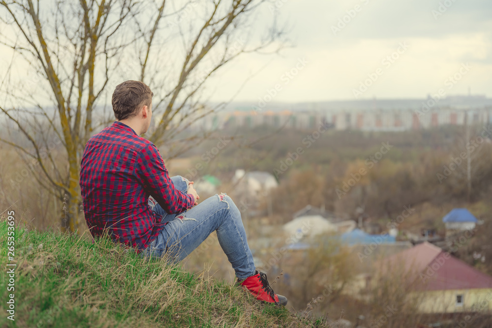 Young man sitting on green hill Side view of young male in jeans and plaid shirt sitting on grass and looking at camera Young man sitting on green hill Side view of young male in jeans and plaid shirt sitting on grass and looking at camera