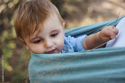 One year old baby boy in a backpack-style sling carried by his mother on a hike in the woods. Modern babywearing in Europe encourages bonding.
