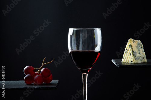 Composition of red wine in glass beside blue cheese and grape on black background.