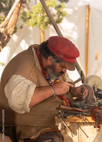 A man dressed as a tinker from the 1800's works on crafting a copper trinket in the shade of a tent.