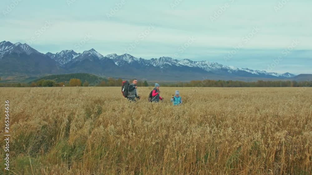 People walk near beautiful mountains in wheat field. Family travels. People environment by mountains, fields. Parents and kids walk using trekking poles. Man and woman have hiking backpacks, flasks. People walk near beautiful mountains in wheat field. Family travels. People environment by mountains, fields. Parents and kids walk using trekking poles. Man and woman have hiking backpacks, flasks.