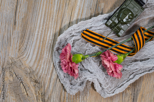 Two pink carnations, Saint George ribbon and military tank on a wooden surface.