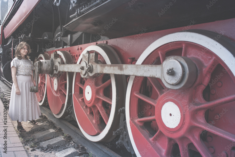 Fototapeta premium Woman stands near the wheels of the locomotive. Girl looks at the big wheel train. Vintage steam train stands on the tracks