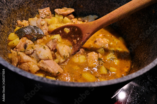 Ragout of potatoes and meat in a saucepan on the stove. A large wooden spoon mixes the lamb stew. Preparation of dishes from potatoes with broth and Bay leaf.