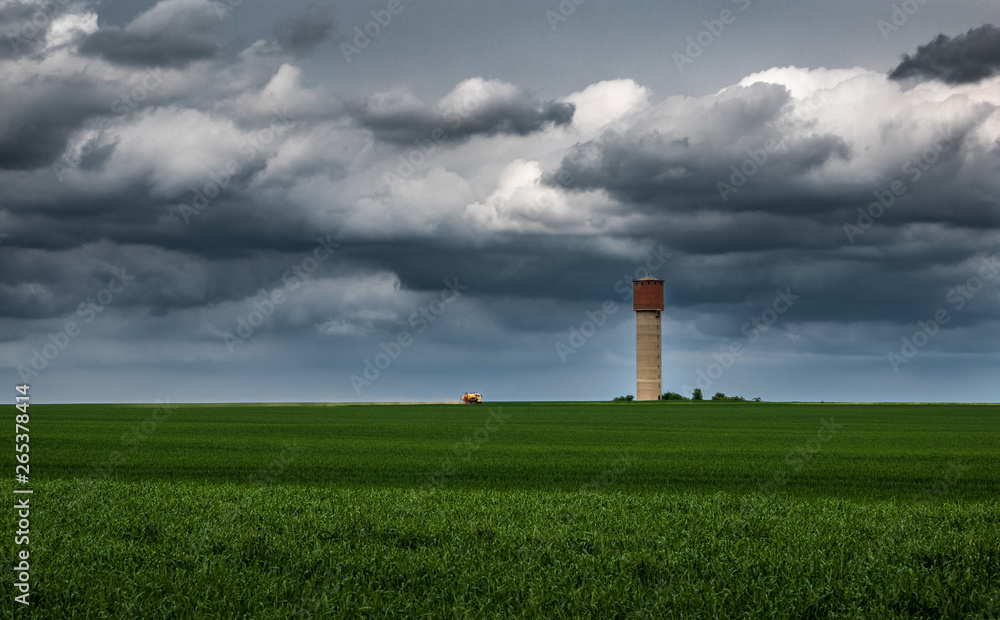 Green field under stormy sky with dark clouds, a water tower and a tank truck in the background