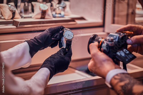 Tattoed photographer is taking photo of expensive watch in posh shop, while shop assistant is holding them.