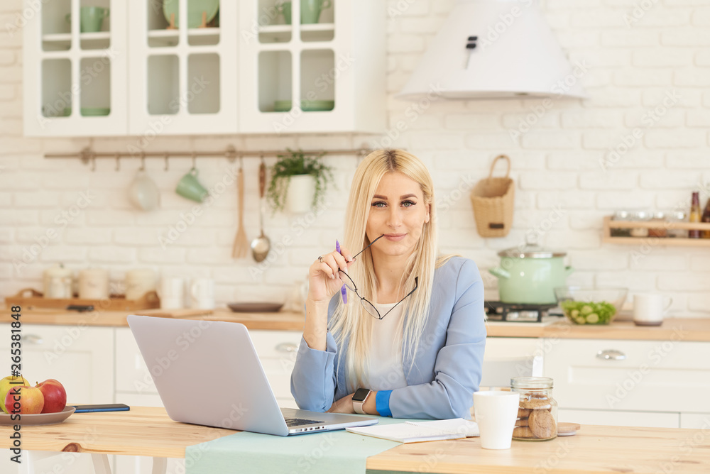 Freelance concept. Beautiful business woman in casual clothes and glasses is examining documents and smiling while working with a laptop in kitchen. Working at home Freelance concept. Beautiful business woman in casual clothes and glasses is examining documents and smiling while working with a laptop in kitchen. Working at home