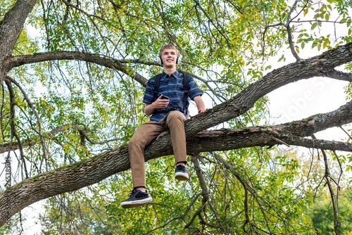 Happy student sitting on a tree branch while listening to music through his headphones.