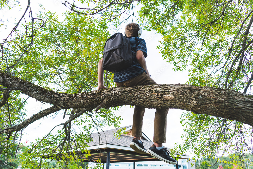 Contemplative student sitting on a tree branch while listening to music through his headphones. Contemplative student sitting on a tree branch while listening to music through his headphones.