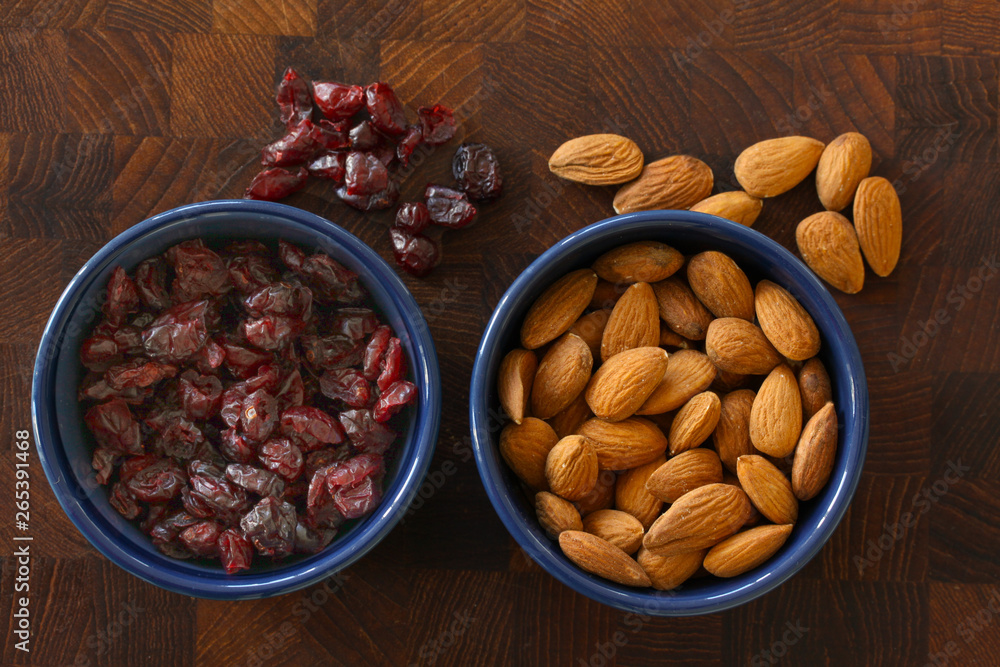 Almonds and cranberries lying in blue bowls on dark wooden background with almonds and cranberries scattered around. Top view image. Almonds and cranberries lying in blue bowls on dark wooden background with almonds and cranberries scattered around. Top view image.