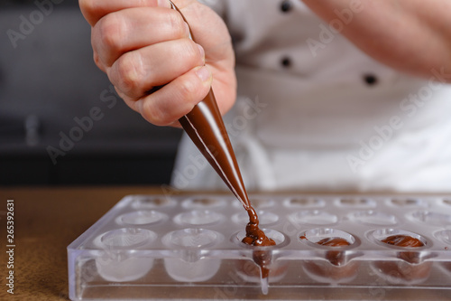 Chocolatier pours chocolate into molds. Chef in white apron using pastry bag filling hot melt chocolate into silicone mold. Concept for making homemade chocolate dessert in modern clean kitchen.